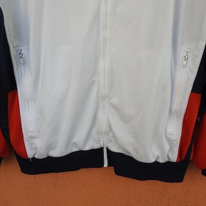 Puede incluir: Una chaqueta deportiva con cremallera que presenta un panel central blanco, paneles laterales azul marino y detalles rojos en las mangas. La prenda incluye dos bolsillos verticales con cremallera en la parte delantera, una cremallera central y un dobladillo acanalado azul marino.