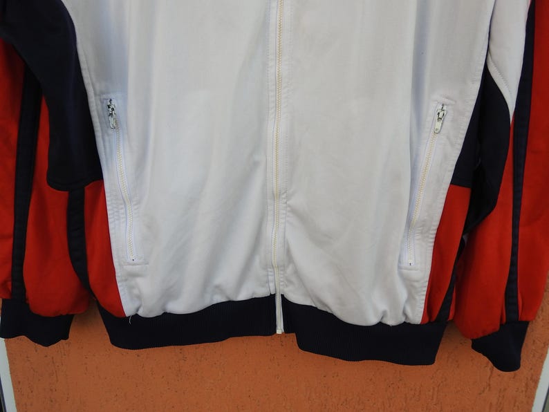 Puede incluir: Una chaqueta deportiva con cremallera que presenta un panel central blanco, paneles laterales azul marino y detalles rojos en las mangas. La prenda incluye dos bolsillos verticales con cremallera en la parte delantera, una cremallera central y un dobladillo acanalado azul marino.