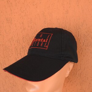 May include: A black baseball cap featuring a red rectangular outline and the red text "THE RENTAL STORE" embroidered on the front panel. The cap has a curved brim with a thin red trim along the edge and a structured crown with a button on top.