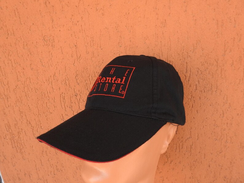 May include: A black baseball cap featuring a red rectangular outline and the red text "THE RENTAL STORE" embroidered on the front panel. The cap has a curved brim with a thin red trim along the edge and a structured crown with a button on top.