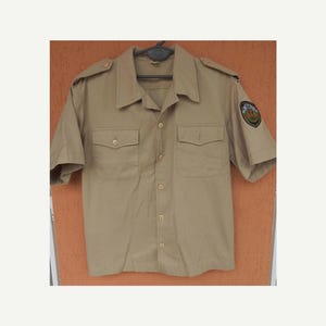 May include: A tan short-sleeved shirt with two chest pockets and shoulder epaulets. The shirt has a button-down front and a patch on the left sleeve. The shirt is hanging on a black hanger.