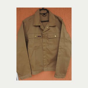 Könnte beinhalten: Eine beigefarbene Arbeitsjacke mit langen Ärmeln, spitzem Kragen, durchgehender Knopfleiste und zwei Brusttaschen mit geknöpften Klappen. Die Jacke ist mit goldfarbenen Metallknöpfen und einem kleinen rechteckigen Markenetikett versehen.