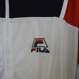 Puede incluir: Una chaqueta deportiva blanca, negra y roja con cierre de cremallera blanca de longitud completa. El pecho muestra un logotipo de FILA bordado en azul marino y rojo. La prenda est&aacute; confeccionada con un material sint&eacute;tico liso y paneles.