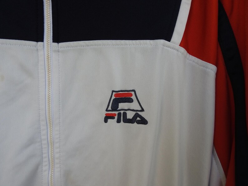 Puede incluir: Una chaqueta deportiva blanca, negra y roja con cierre de cremallera blanca de longitud completa. El pecho muestra un logotipo de FILA bordado en azul marino y rojo. La prenda est&aacute; confeccionada con un material sint&eacute;tico liso y paneles.