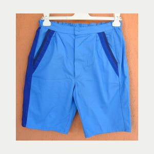 May include: Light blue shorts with dark blue accents on the sides and pockets. The shorts are hanging on a white hanger against an orange background. The shorts have a drawstring waistband.
