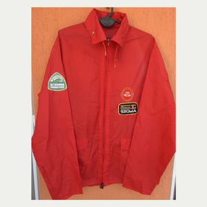 May include: A bright red jacket with a zip-up front, collar, and two front pockets. The jacket features three patches: one with the text "Shakespeare", one with a circular logo, and one with the text "SIGMA".