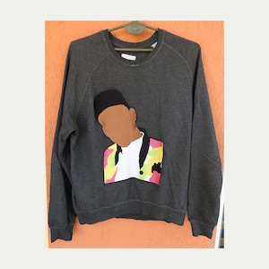 Op de afbeelding: Donkergrijs sweatshirt met een kleurrijke afbeelding van een persoon met een zwarte hoed en een jasje met patroon. De afbeelding staat op de voorkant van het sweatshirt, dat aan een groene hanger hangt.