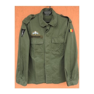 May include: An olive green military-style jacket featuring a collar, buttoned shoulder epaulets, and two chest pockets. The right sleeve has a black patch with a white sword, while the left sleeve displays a small Belgian flag patch.