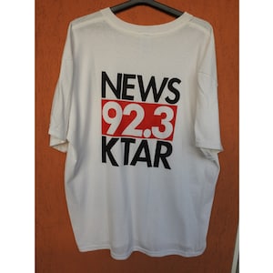 Può includere: Maglietta bianca con testo nero che dice "NEWS 92.3 KTAR" in un riquadro rosso.