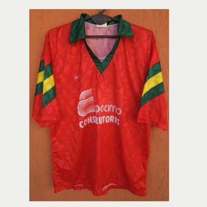 May include: A red short-sleeved jersey with a V-neck collar in dark green. The sleeves feature yellow and green diagonal stripes. The front displays the white text "Espacimo CONSTRUTORES" and a stylized logo above the brand name.