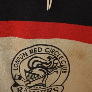 Pode incluir: Close-up de uma camisola de h&oacute;quei vintage. A camisola apresenta um esquema de cores preto, vermelho e creme. As palavras "LONDON RED CIRCLE CLUB" s&atilde;o impressas acima de um log&oacute;tipo de capacete de cavaleiro. A palavra "RAIDERS" &eacute; impressa abaixo.