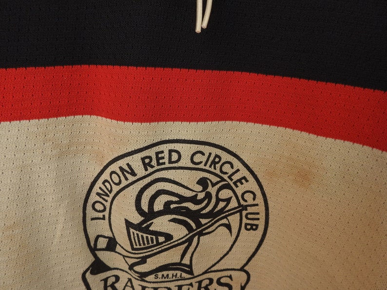 Pode incluir: Close-up de uma camisola de h&oacute;quei vintage. A camisola apresenta um esquema de cores preto, vermelho e creme. As palavras "LONDON RED CIRCLE CLUB" s&atilde;o impressas acima de um log&oacute;tipo de capacete de cavaleiro. A palavra "RAIDERS" &eacute; impressa abaixo.