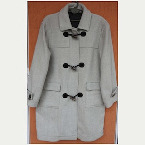Peut inclure: Un duffle-coat gris clair avec un col et trois fermetures à bascule. Le manteau a deux poches avant et des poignets boutonnés. Le manteau est suspendu à un cintre.