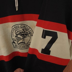 Pode incluir: Camisola de h&oacute;quei preta, vermelha e creme com o texto "LONDON RED CIRCLE CLUB RAIDERS" e o n&uacute;mero 7 na manga. A camisola tem um design de malha e um cord&atilde;o no pesco&ccedil;o.