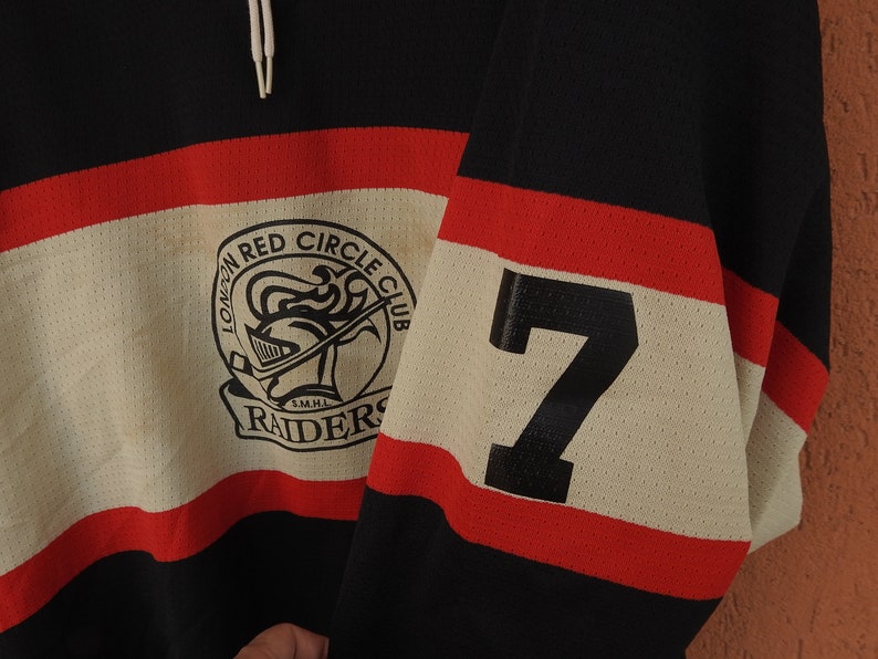 Pode incluir: Camisola de h&oacute;quei preta, vermelha e creme com o texto "LONDON RED CIRCLE CLUB RAIDERS" e o n&uacute;mero 7 na manga. A camisola tem um design de malha e um cord&atilde;o no pesco&ccedil;o.