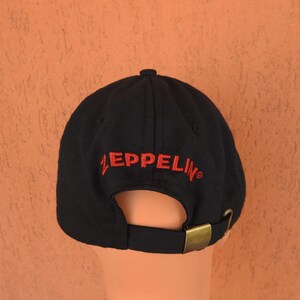May include: A black baseball cap featuring the word ZEPPELIN embroidered in red thread above the rear opening. The cap includes a fabric strap with an adjustable antique brass-toned metal buckle closure at the back for a secure and customizable fit.