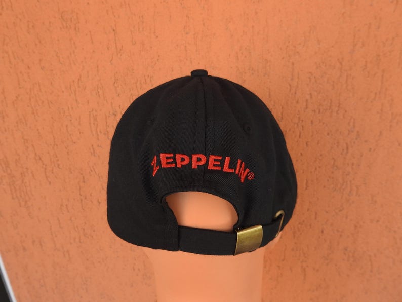 May include: A black baseball cap featuring the word ZEPPELIN embroidered in red thread above the rear opening. The cap includes a fabric strap with an adjustable antique brass-toned metal buckle closure at the back for a secure and customizable fit.