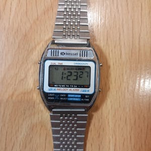Op de afbeelding: Een zilverkleurig Kessel digitaal horloge met een rechthoekige wijzerplaat met de tijd 1:23:27. Het horloge heeft functies voor dubbele tijd, chronograaf en melodie-alarm. De horlogeband is gemaakt van bijpassend zilverkleurig metaal.