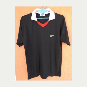 Op de afbeelding: Zwart T-shirt met korte mouwen, een witte kraag en rode bies. Het shirt heeft een V-hals en het Reebok-logo is geborduurd op de voorkant.