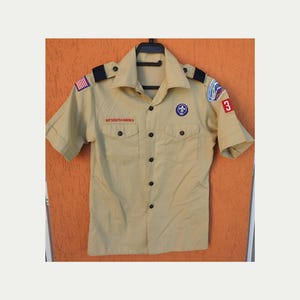 May include: Tan short-sleeved Boy Scouts of America uniform shirt. The shirt has a button-down front, two chest pockets, and patches on the sleeves. The patches include the American flag, a Boy Scout emblem, and a number 3.