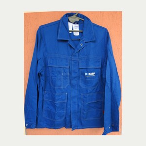 May include: A bright blue long-sleeve work jacket featuring a classic collar, a button-up front, and four large front pockets with flaps. The right chest pocket displays the white BASF logo and text. The jacket is displayed on a plastic hanger.