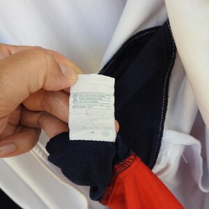 Puede incluir: Una prenda de color blanco, azul marino y rojo con una etiqueta de cuidado interior que incluye las instrucciones impresas MACHINE WASH WARM, NON LANDED CLAIRE, DO NOT BLEACH y WARM IRON. La etiqueta blanca tiene texto negro y s&iacute;mbolos.