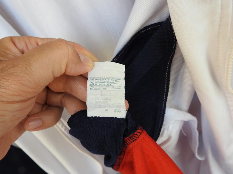 Puede incluir: Una prenda de color blanco, azul marino y rojo con una etiqueta de cuidado interior que incluye las instrucciones impresas MACHINE WASH WARM, NON LANDED CLAIRE, DO NOT BLEACH y WARM IRON. La etiqueta blanca tiene texto negro y s&iacute;mbolos.