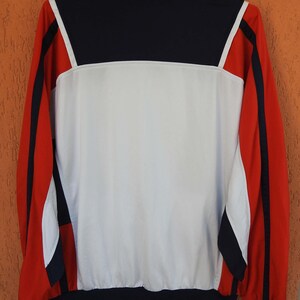 Puede incluir: Una chaqueta deportiva de manga larga con un dise&ntilde;o de bloques de color en azul marino, blanco y rojo. El panel trasero es principalmente blanco con una secci&oacute;n superior azul marino, mientras que las mangas son rojas con rayas azul marino.
