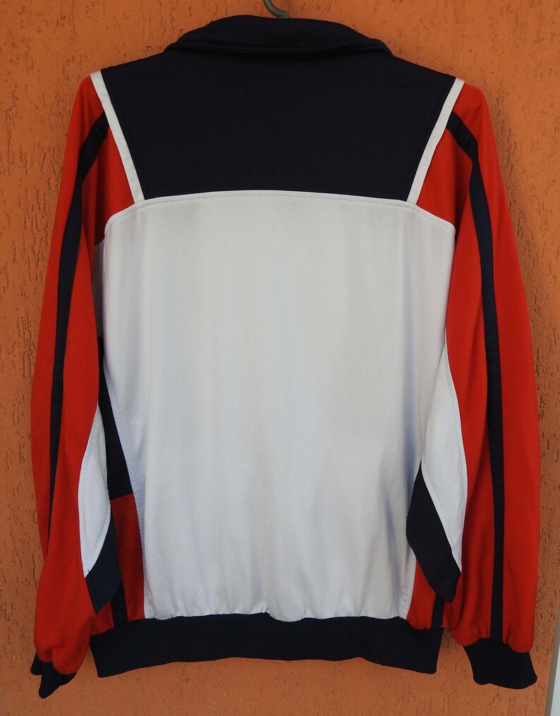 Puede incluir: Una chaqueta deportiva de manga larga con un dise&ntilde;o de bloques de color en azul marino, blanco y rojo. El panel trasero es principalmente blanco con una secci&oacute;n superior azul marino, mientras que las mangas son rojas con rayas azul marino.