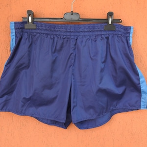 Puede incluir: Pantalones cortos deportivos azul marino con rayas laterales azul claro. Los pantalones cortos están hechos de un material brillante y ligero y tienen una cintura elástica. Cuelgan de una percha negra sobre un fondo naranja.