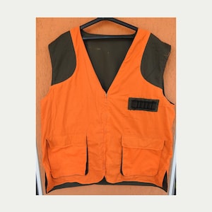 Op de afbeelding: Een mouwloos vest met een feloranje romp en donker olijfgroene schouderpanelen. Het kledingstuk is voorzien van een centrale ritssluiting, twee grote vierkante voorzakken en een kleine horizontale zwarte strook met lussen op de borst.