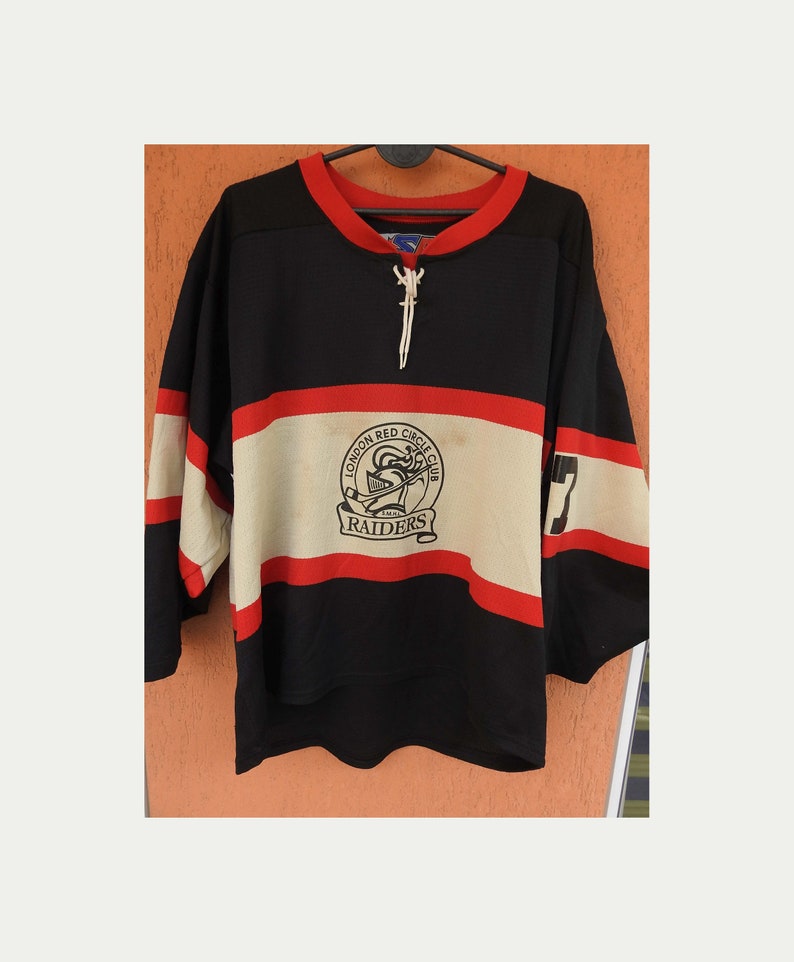 Pode incluir: Camisola de h&oacute;quei preta com detalhes em vermelho e creme. A camisola apresenta o texto "LONDON RED CIRCLE CLUB RAIDERS" e o n&uacute;mero "7" na manga. Possui gola com atacadores e est&aacute; pendurada num cabide preto.