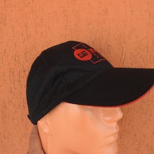 May include: A black baseball cap featuring a red embroidered CAT logo inside a rectangular frame on the front panel. The cap has a curved brim with a thin red trim along the edge, providing a structured look with a classic six-panel design.