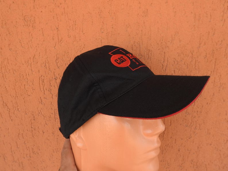 May include: A black baseball cap featuring a red embroidered CAT logo inside a rectangular frame on the front panel. The cap has a curved brim with a thin red trim along the edge, providing a structured look with a classic six-panel design.
