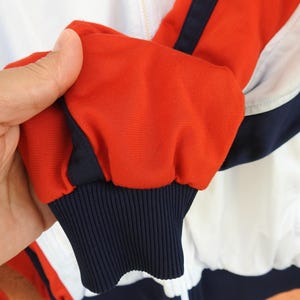 Puede incluir: Una chaqueta deportiva con dise&ntilde;o de bloques de color en rojo vibrante, blanco n&iacute;tido y azul marino profundo. La manga termina en un pu&ntilde;o acanalado de color azul marino y la prenda incluye una cremallera frontal completa con riel blanco.