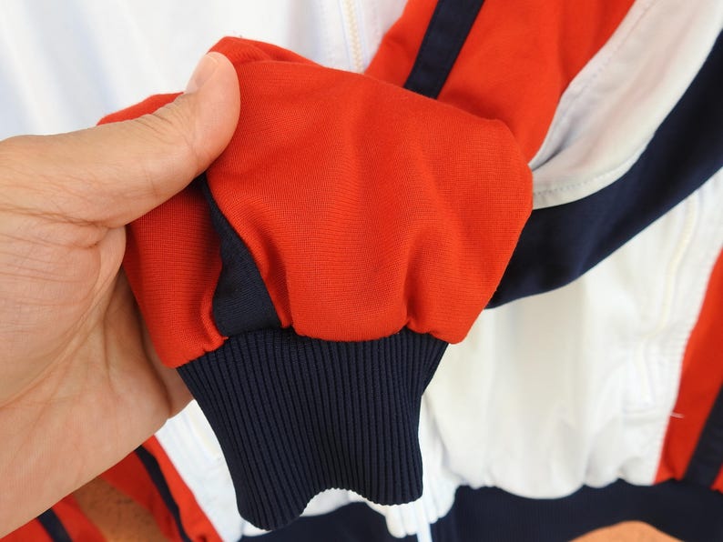 Puede incluir: Una chaqueta deportiva con dise&ntilde;o de bloques de color en rojo vibrante, blanco n&iacute;tido y azul marino profundo. La manga termina en un pu&ntilde;o acanalado de color azul marino y la prenda incluye una cremallera frontal completa con riel blanco.
