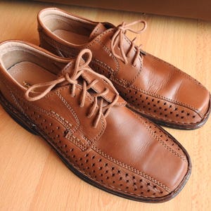 May include: A pair of brown leather dress shoes with a lace-up design. The shoes have a perforated pattern on the toe and sides. The shoes are sitting on a wooden surface.