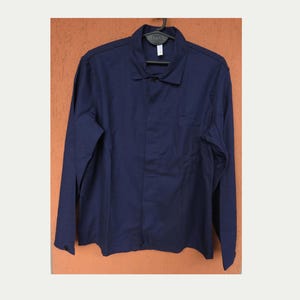 Könnte beinhalten: Eine einfarbige, marineblaue Jacke mit langen Ärmeln, klassischem Kragen, einer Brusttasche auf der linken Seite und einer Knopfleiste vorne. Das Kleidungsstück hängt auf einem schwarzen Kleiderbügel vor einem strukturierten orangefarbenen Hintergrund.