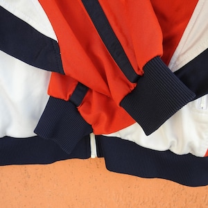 Puede incluir: Una chaqueta deportiva con cremallera que presenta un dise&ntilde;o de bloques de color en rojo vibrante, blanco n&iacute;tido y azul marino intenso. La prenda incluye mangas largas con pu&ntilde;os acanalados en azul marino, un dobladillo acanalado a juego y una cremallera central blanca.