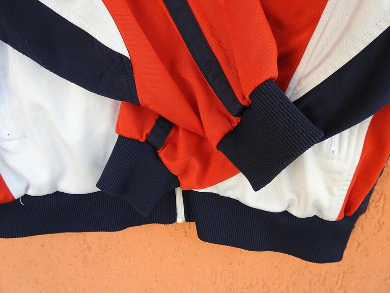 Puede incluir: Una chaqueta deportiva con cremallera que presenta un dise&ntilde;o de bloques de color en rojo vibrante, blanco n&iacute;tido y azul marino intenso. La prenda incluye mangas largas con pu&ntilde;os acanalados en azul marino, un dobladillo acanalado a juego y una cremallera central blanca.