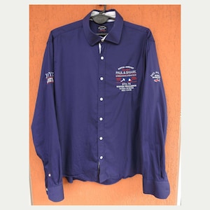May include: A long-sleeve navy blue button-down shirt featuring white embroidery on the chest that reads "BOSTON-NEWPORT YACHT PAUL & SHARK INTERNATIONAL OCEAN TEAM NYL 74 WINTER CHALLENGE ROYAL NORTH SEA CLUB" with small red and white flag icons.