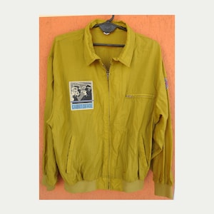 May include: A mustard yellow bomber jacket with a zipper closure and a patch on the left chest that says "CARNET DE VOL".