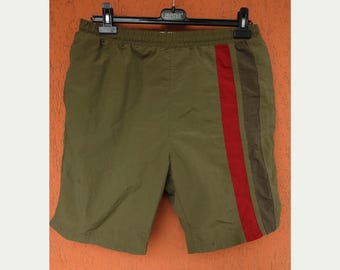 May include: Olive green shorts with a vertical stripe in red, brown, and olive green. The shorts are hanging on a black hanger. The shorts appear to be made of a lightweight, possibly nylon, material.