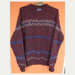 Puede incluir: Un suéter granate con cuello redondo y mangas largas. El suéter presenta un patrón geométrico en azul, gris y blanco en el pecho y las mangas. La tela tiene una apariencia texturizada.