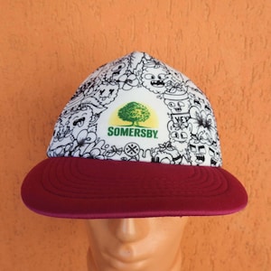 Könnte beinhalten: Eine Baseballkappe mit weißer Krone, einem schwarz-weißen Kritzelmuster und einem zentralen Somersby-Logo mit einem grünen Baum. Die Kappe hat einen einfarbigen kastanienbraunen Schirm mit horizontalen Ziernähten auf der Oberfläche.