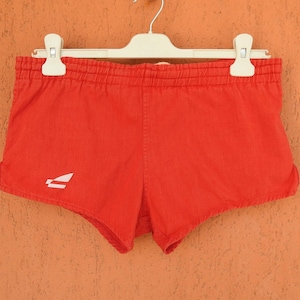 May include: Bright red athletic shorts featuring an elasticized waistband and a small white graphic logo on the lower left side. The shorts are displayed on a white plastic hanger, showcasing a simple, functional design with short leg openings.