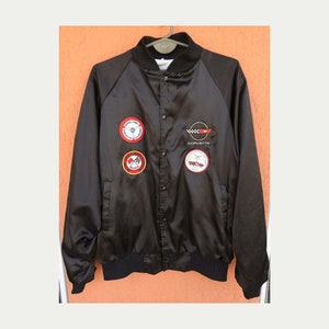 May include: Black satin bomber jacket with multiple embroidered patches, including a Corvette logo, a checkered flag, and a car racing scene.