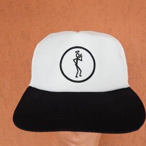 Op de afbeelding: Een wit en zwarte baseballcap met een zwarte klep. De cap heeft een cirkelvormig embleem met een stokfiguur met een halo en een sigaret. De achtergrond is effen oranje.