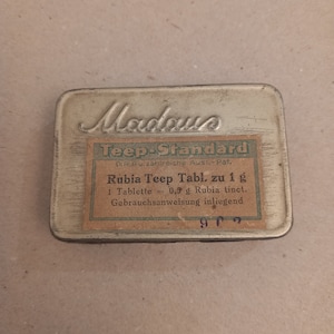 Puede incluir: Una lata de metal plateada con una etiqueta que dice "Madawa Teep-Standard D.R.P. zahlreiche Ausi.-Pat. Rubia Teep Tabl. zu 1 g 1 Tablette = 0,5 g Rubia tinct. Gebrauchsanweisung inliegend"