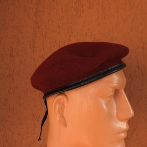 May include: A burgundy beret with a black leather band is displayed on a mannequin head. The beret is made of a soft, felt-like material and has a classic, round shape. The background is a textured, orange-brown wall.
